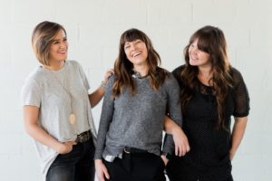 3 women smiling and standing beside white wall