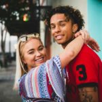 smiling woman with both hands wrap around man's neck near white concrete building